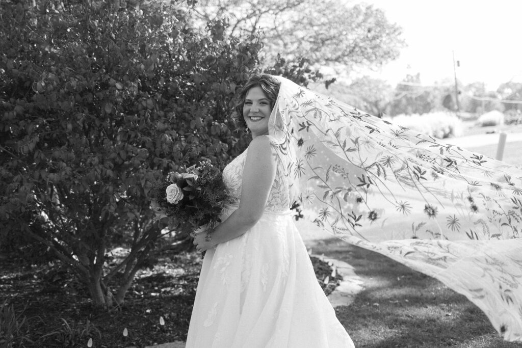 A bride in a white dress smiles while standing outdoors. Her veil is decorated with floral patterns and flows elegantly in the breeze. She holds a bouquet of flowers, and trees are visible in the background. The scene is captured in black and white.