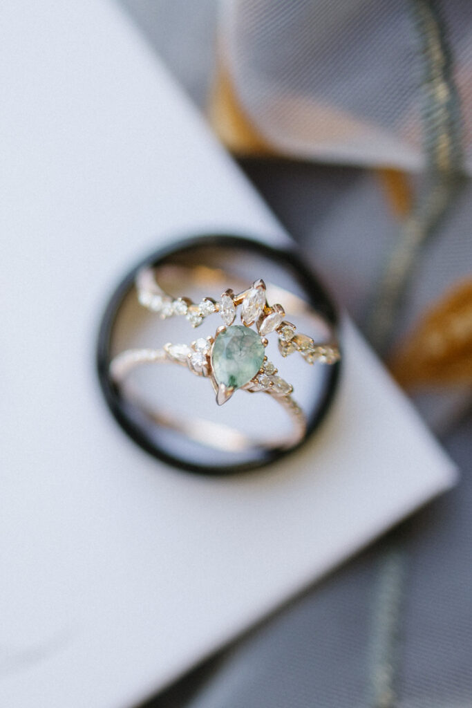 A close-up of a gold ring featuring a central light green gemstone surrounded by smaller clear stones. The ring rests on a black band atop a gray and beige patterned fabric and a white surface.