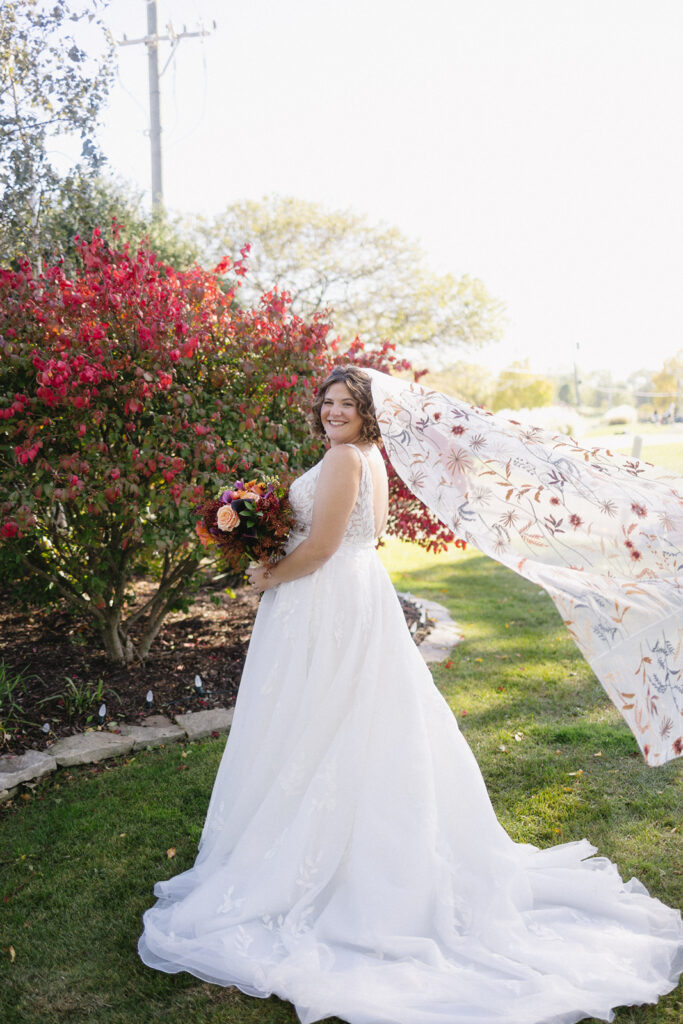 A bride in a flowing white wedding dress stands outdoors near a red-leaved bush. She holds a bouquet and smiles at the camera. Her long veil is caught in the breeze, creating a sense of movement. The scene is bright and sunny.