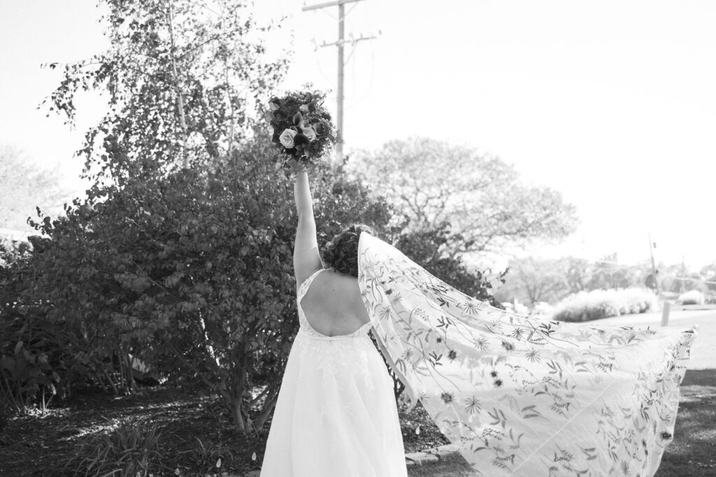 Bride with flowing floral veil raises bouquet triumphantly, standing outdoors near lush greenery and trees. Back view in black and white.