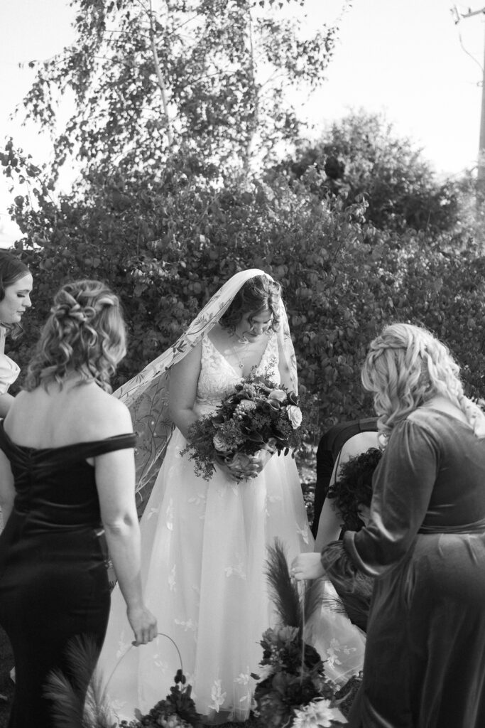 A bride in an elegant gown stands holding a bouquet, surrounded by bridesmaids outdoors. She wears a long veil, and the scene is set against lush foliage. The image is in black and white, capturing a serene, intimate moment.