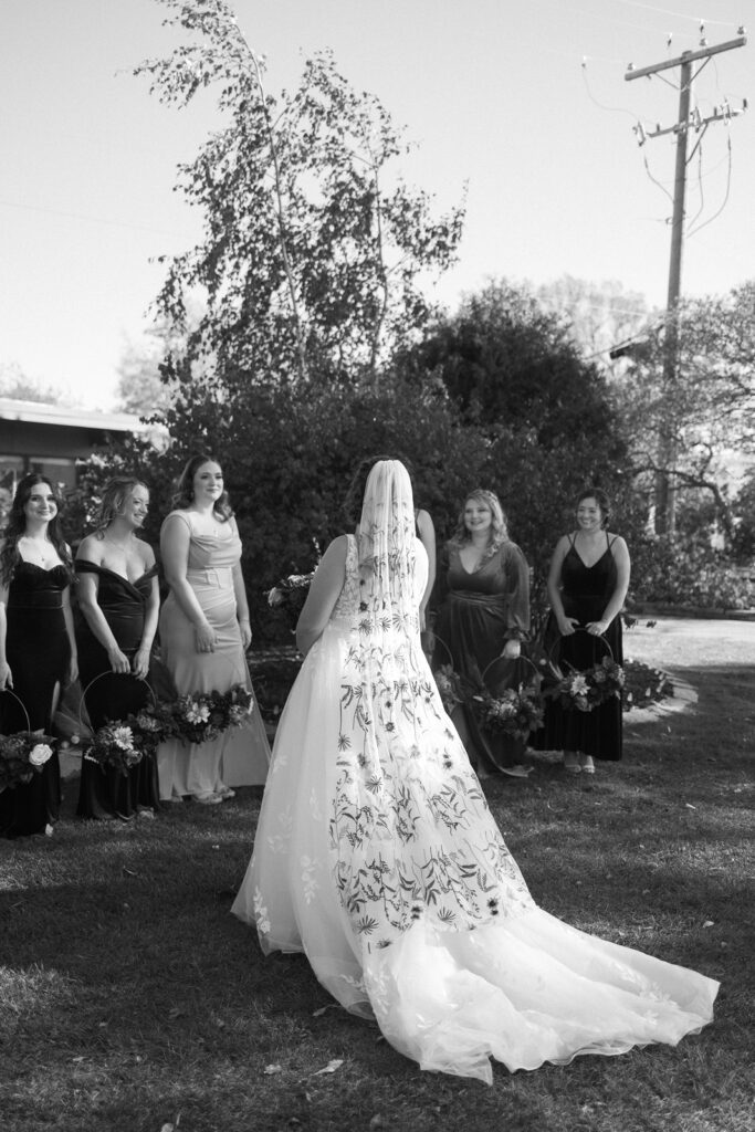 Bride in a flowing gown with a long veil stands with bridesmaids outdoors. Bridesmaids wear dark dresses and hold bouquets. Trees and a power pole are visible in the background under a clear sky. Image is in black and white.