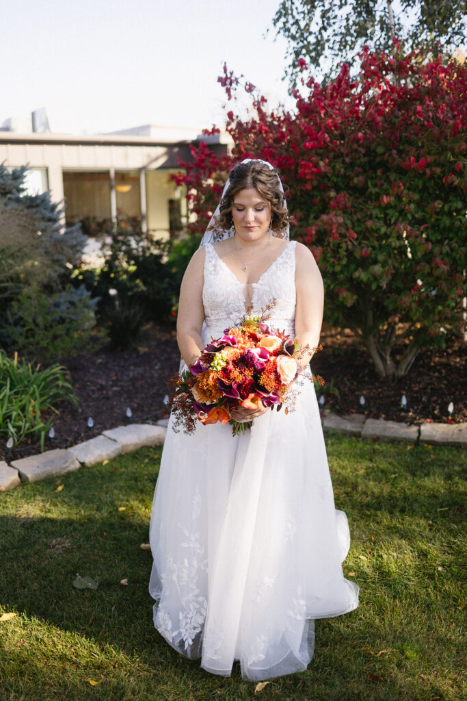 A bride in a white wedding dress holds a vibrant bouquet of flowers, standing on a lawn with colorful red and green foliage in the background. She looks down at the bouquet, under a sunny, clear sky.