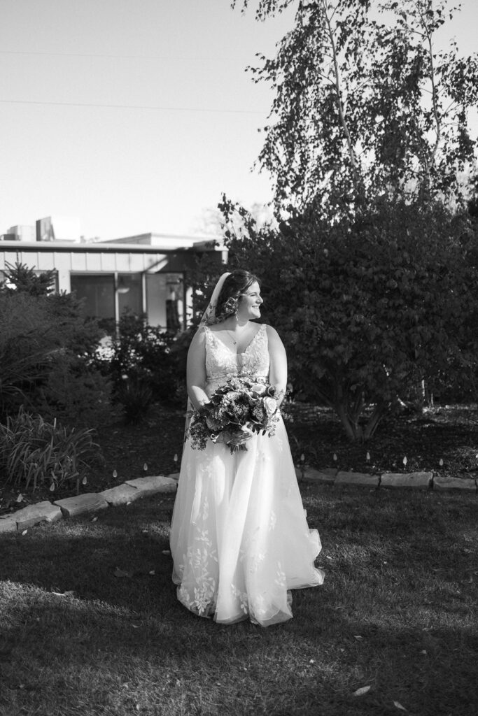 Bride in a wedding dress holds a bouquet and stands on grass near landscaped bushes. Shes looking to the side under a tree with sunlight casting shadows. The setting includes a building in the background. The photo is in black and white.