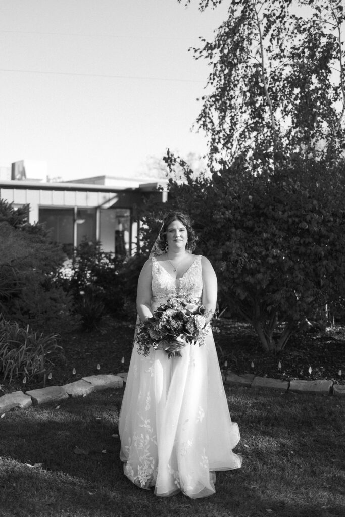 A bride stands outdoors holding a bouquet. She is wearing a long, flowing white wedding dress with lace details. The background features trees and a building. The image is in black and white.