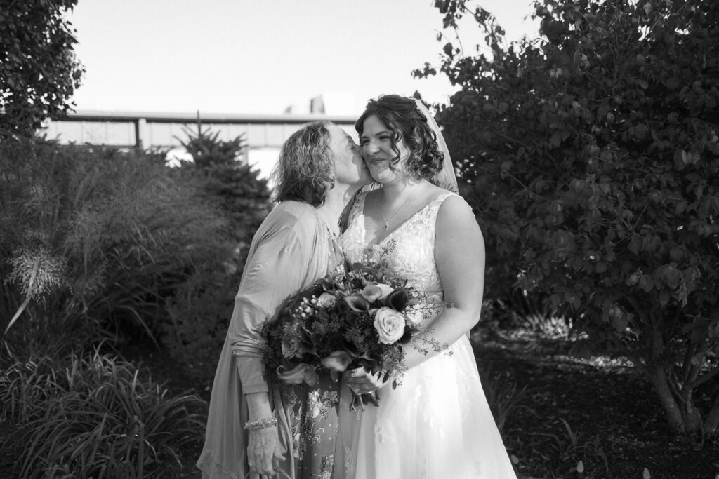 A bride in a white dress holds a bouquet and smiles as an older woman kisses her on the cheek. They are standing outdoors, surrounded by greenery and trees, with a building in the background. The image is in black and white.