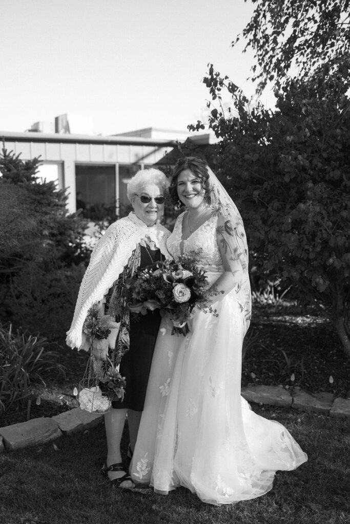 A black and white photo of a bride in a wedding dress holding a bouquet and standing next to an older woman in sunglasses and a shawl. They are outdoors, with trees and a building in the background. Both are smiling at the camera.