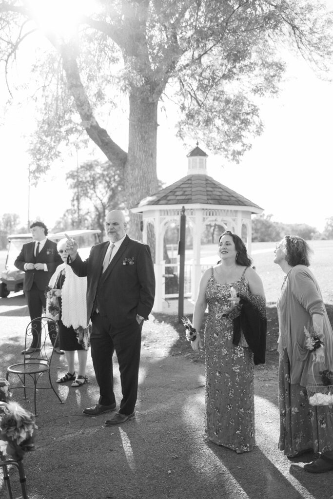 A black and white photo of a group of people outdoors near a gazebo. They are dressed in formal attire, with one person in a suit and others in dresses. The sunlight filters through the trees, creating a warm, inviting atmosphere.