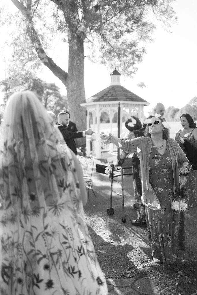 A black-and-white photo of a wedding scene. A person in a veil faces a woman with sunglasses throwing flower petals. A gazebo and other guests are visible in the background, with trees and sunlight filtering through.