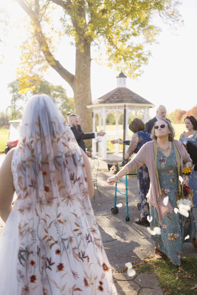 A bride in a floral-patterned dress approaches guests at an outdoor wedding, with a gazebo in the background. The guests, including a woman with a walker and sunglasses, appear joyful, holding white petals. Its a sunny day with green and yellow foliage.