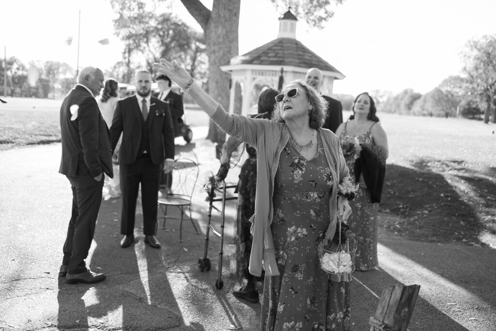 A woman in sunglasses waves her arm enthusiastically, holding a small bag. She is surrounded by people dressed formally, including a man with a cane. A gazebo and trees are in the background under bright sunlight. The scene conveys a joyful gathering.