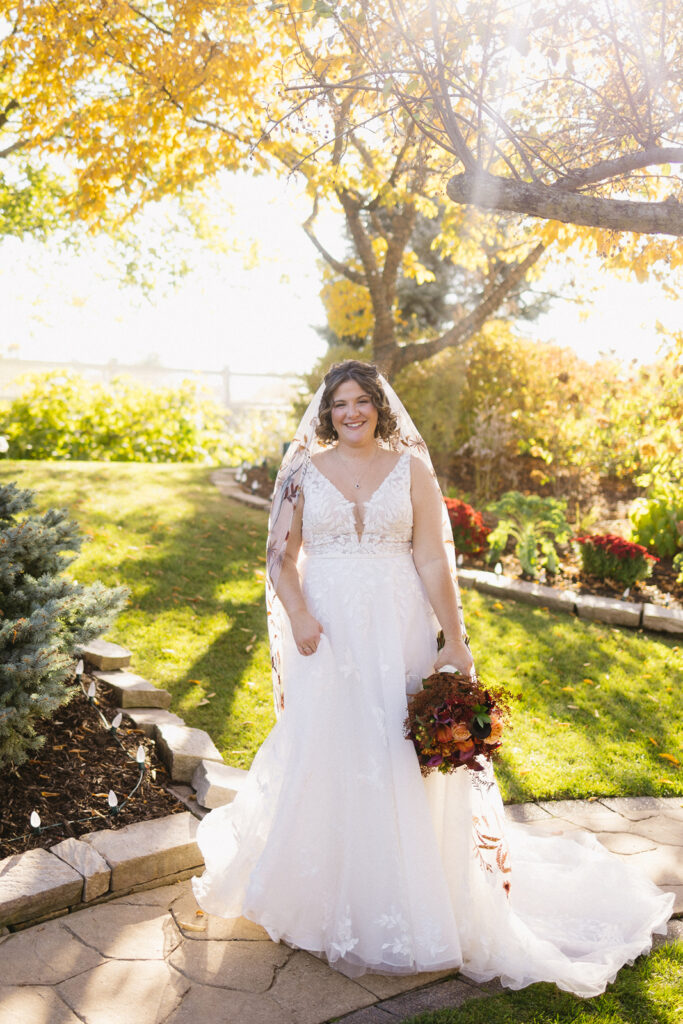 A bride in a white gown smiles under a tree with golden leaves. She holds a bouquet of red and orange flowers and wears a veil with leaf patterns. Sunlight filters through the tree, creating a warm, bright atmosphere. A garden path is visible in the background.