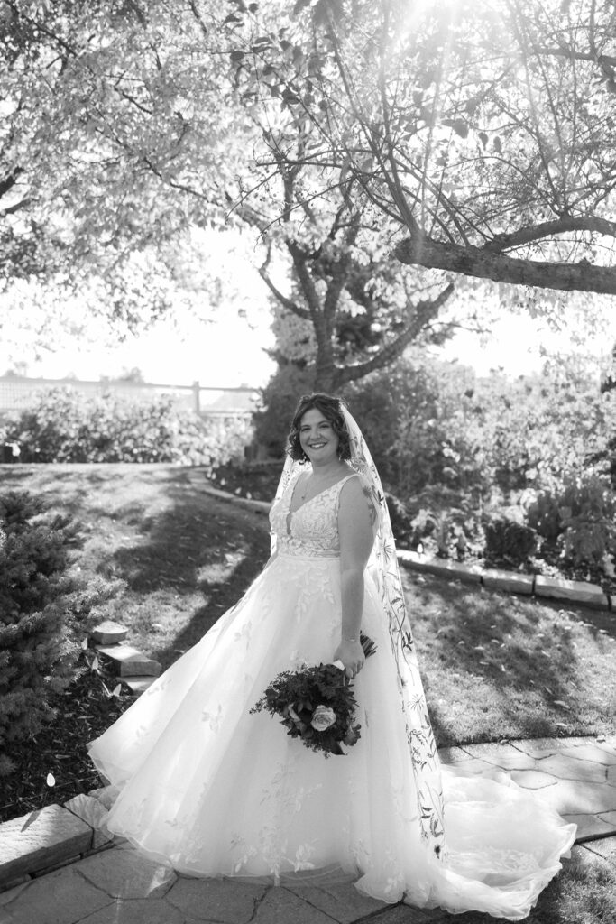 A bride in a white gown and veil stands on a garden path, holding a bouquet. Sunlight filters through the trees above, casting soft shadows. The scene is serene and picturesque.