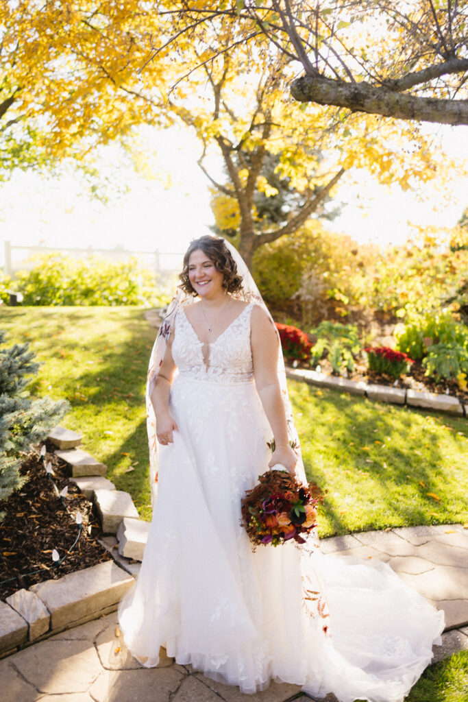 Bride in a white gown and veil stands outdoors on a sunny day, holding a bouquet of autumn flowers. She is smiling, surrounded by trees with golden leaves and lush greenery in a garden setting.