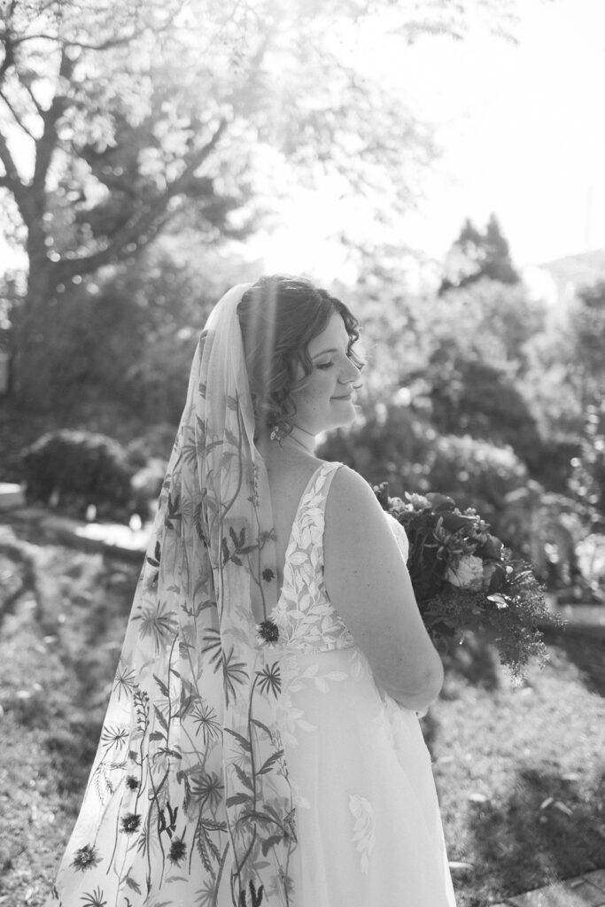 A bride in a white dress stands outside, smiling with eyes closed. She holds a bouquet and wears a veil adorned with floral patterns. Sunlight filters through trees, creating a serene, joyful atmosphere. Image is in black and white.