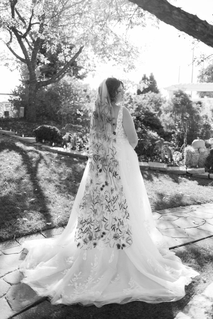 Black and white image of a bride standing outdoors on a stone path, wearing a long, flowing gown with floral embroidery. Her back is turned, and she faces a garden with trees and sunlight filtering through the branches.