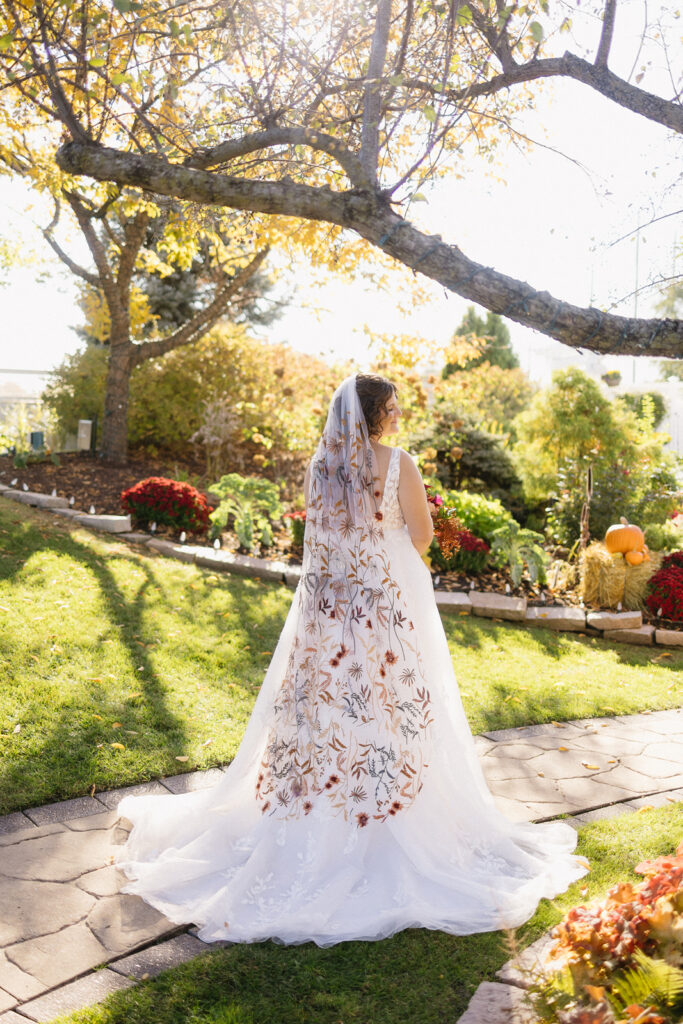 Bride in a white wedding dress with a colorful floral-patterned veil stands in a sunlit garden. Surroundings include a stone path, lush green grass, autumn trees, and pumpkins, creating a warm, serene atmosphere.