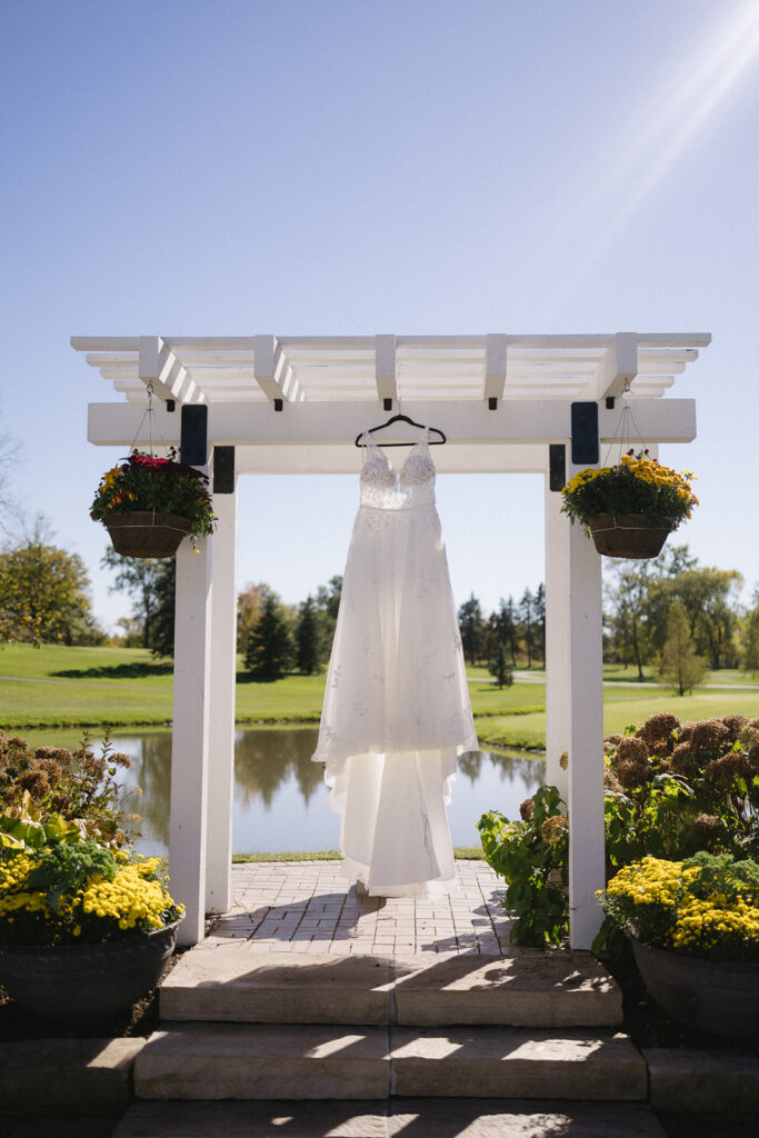 A white wedding dress hangs on a wooden pergola by a pond. Potted flowers adorn the structure, and trees are visible in the bright sunlight in the background, creating a serene outdoor setting.