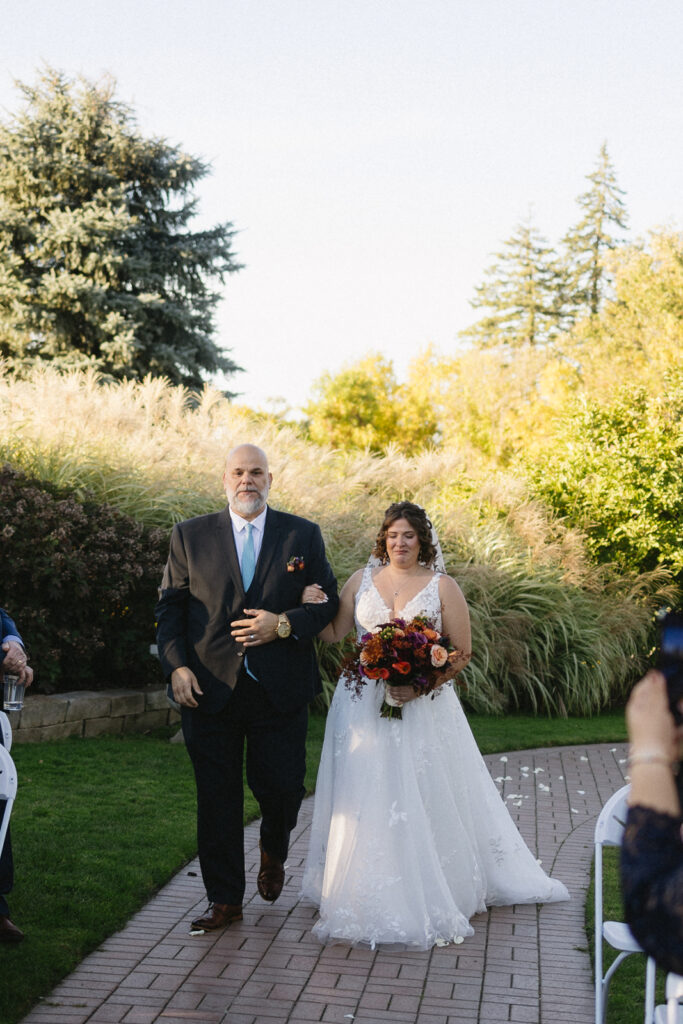 A bride in a white gown holding a bouquet walks down a brick path with an older man in a dark suit. They are surrounded by greenery and grasses, under a clear blue sky, with seated guests watching.