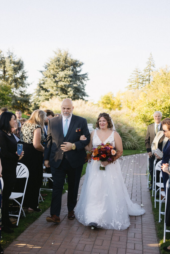 A bride in a white dress holding a bouquet of colorful flowers walks down a brick path with a man in a suit. They are surrounded by seated guests outdoors on a sunny day. Trees and greenery are visible in the background.