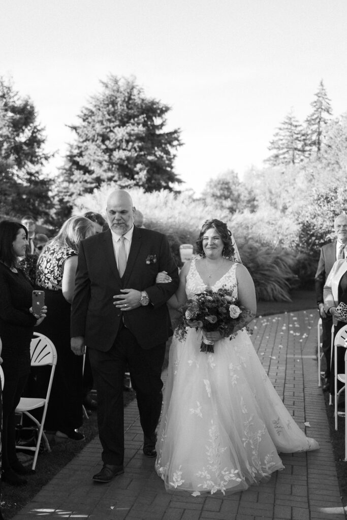 A bride in a white gown holds a bouquet while walking down an outdoor aisle with an older man in a suit. Guests are seated on either side, and trees are visible in the background. The scene is in black and white.