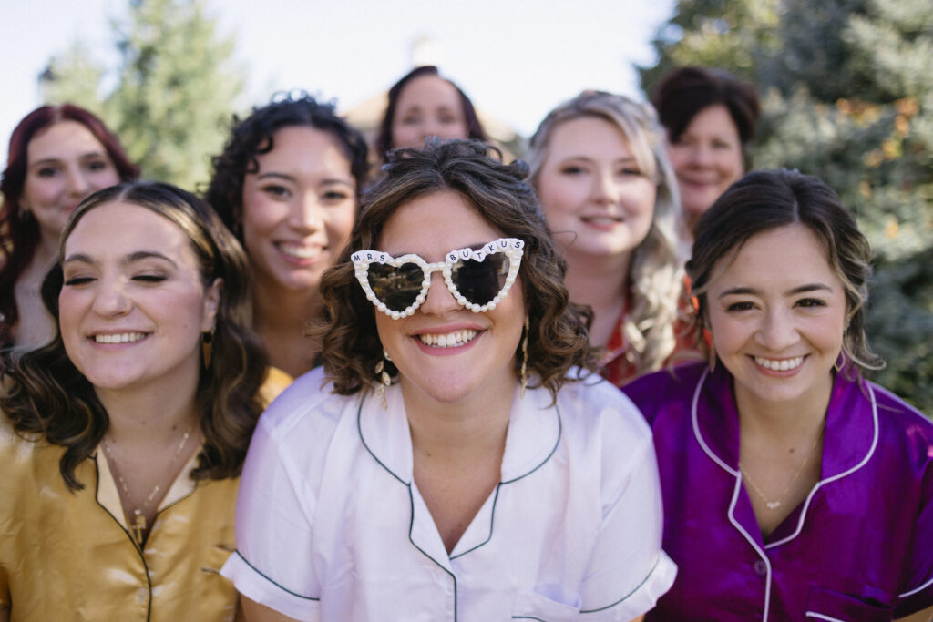 A group of smiling women, some wearing colorful pajamas, stand together outdoors. One woman in front wears heart-shaped sunglasses. Trees are visible in the background.