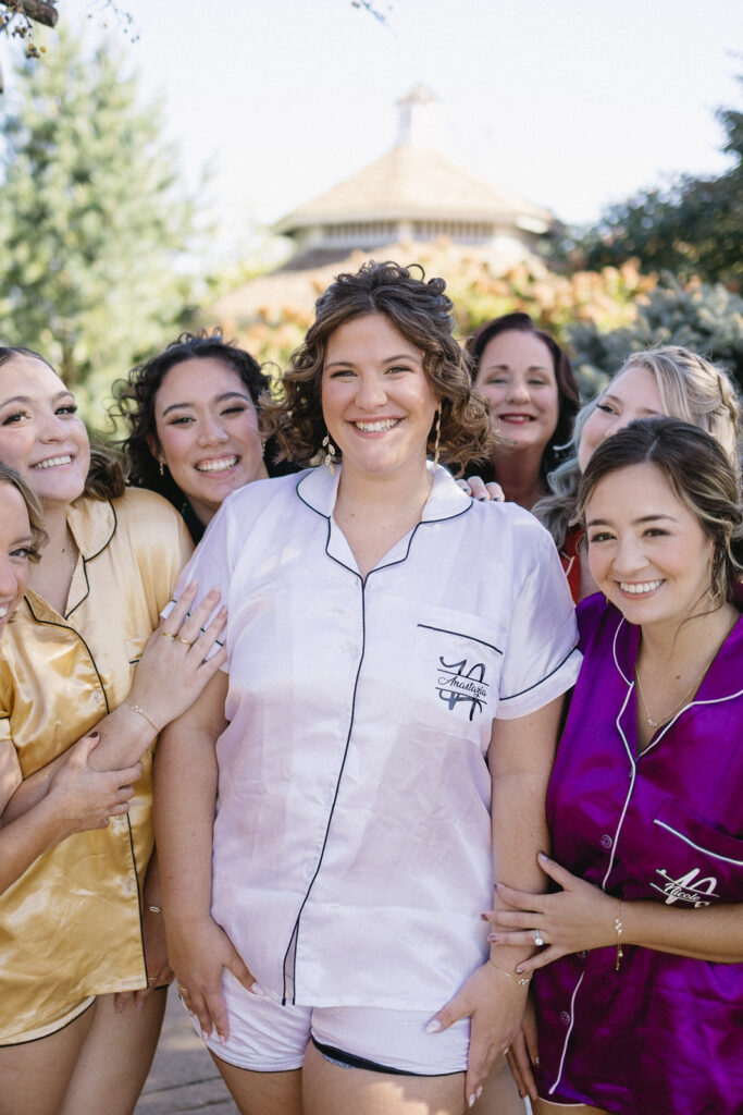 A joyful group of women in silk pajamas stands outdoors. The central woman in white is smiling, surrounded by friends in colorful pajamas: yellow, pink, and purple. The background features trees and a blurred building.