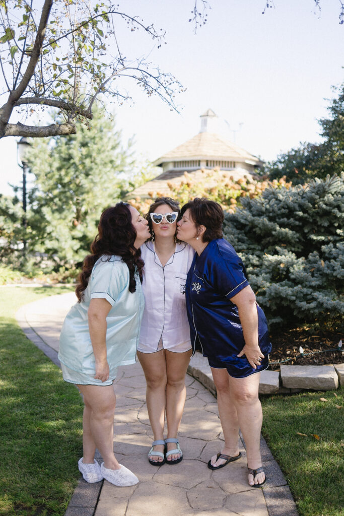 Three women stand on a garden path, wearing matching pajamas. The central woman, in white pajamas and heart-shaped sunglasses, is being kissed on both cheeks by the others in blue and teal pajamas. Trees and shrubs surround them.