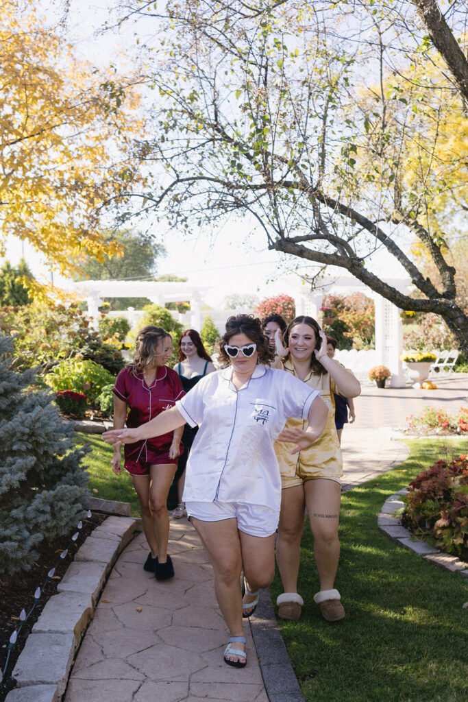 A group of people joyfully walks outside in pajamas. The foreground features a person in white pajamas and heart-shaped sunglasses, with others in colorful pajamas behind. They are surrounded by a garden with autumn trees and bright sunlight.