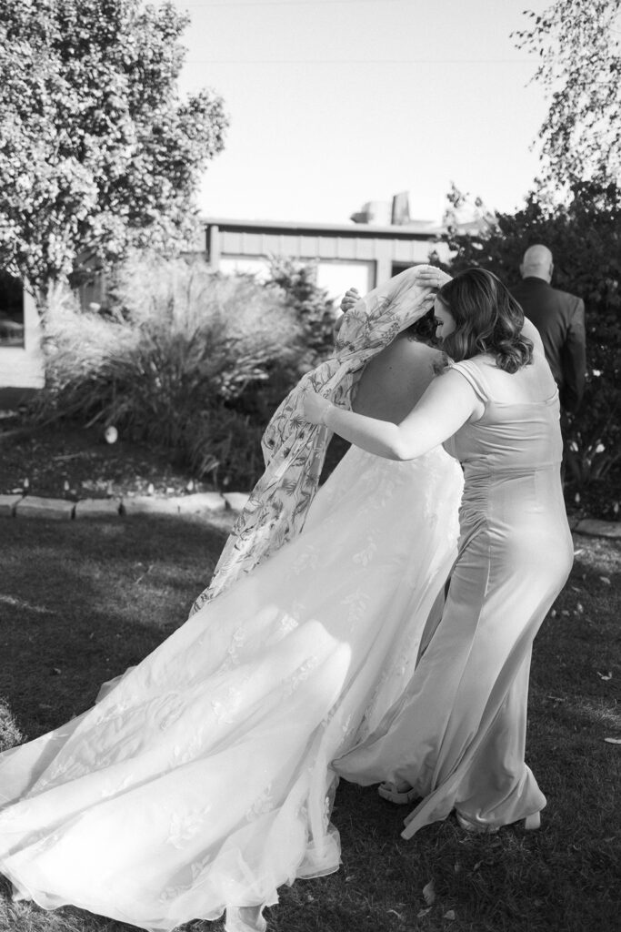 A black and white photo of two women in elegant dresses embracing outdoors. One wears a long, flowing gown and a veil, while the other has a fitted dress. They stand on grass with trees and a building in the background.