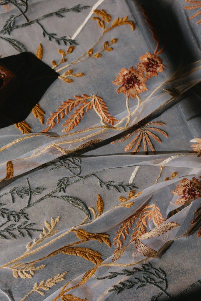 Close-up of sheer fabric with intricate floral embroidery in shades of orange, gold, and green. The fabric is gently draped, with sunlight casting soft shadows across its surface.