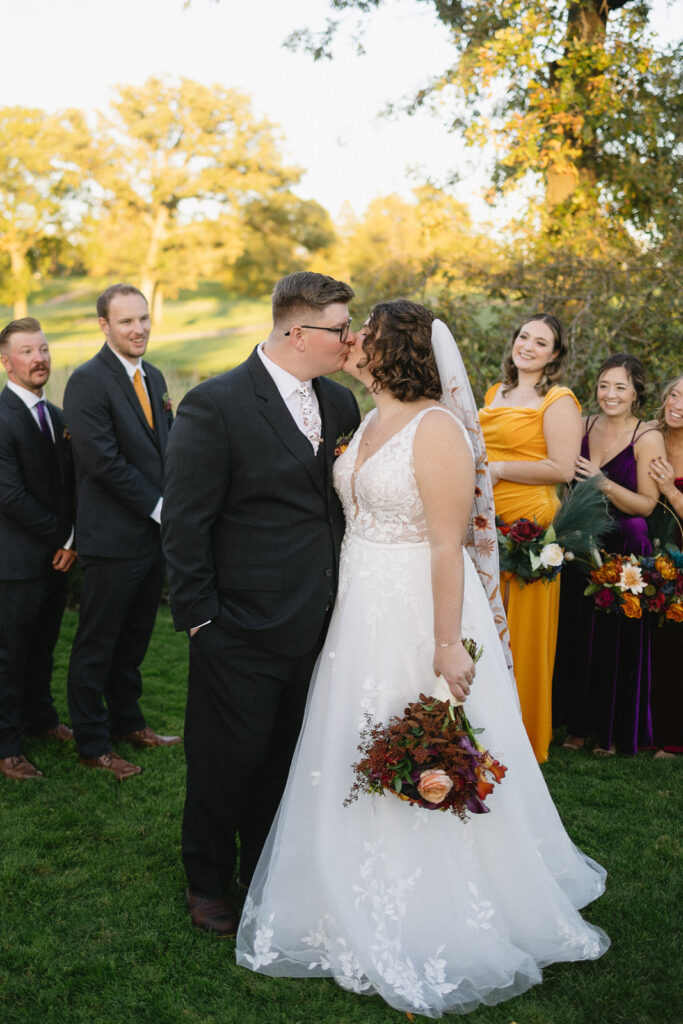 A bride and groom share a kiss outdoors, surrounded by their smiling wedding party. The bride holds a bouquet of flowers. The bridesmaids wear vibrant dresses, and the groomsmen are in suits. The setting is sunny with trees and grass.