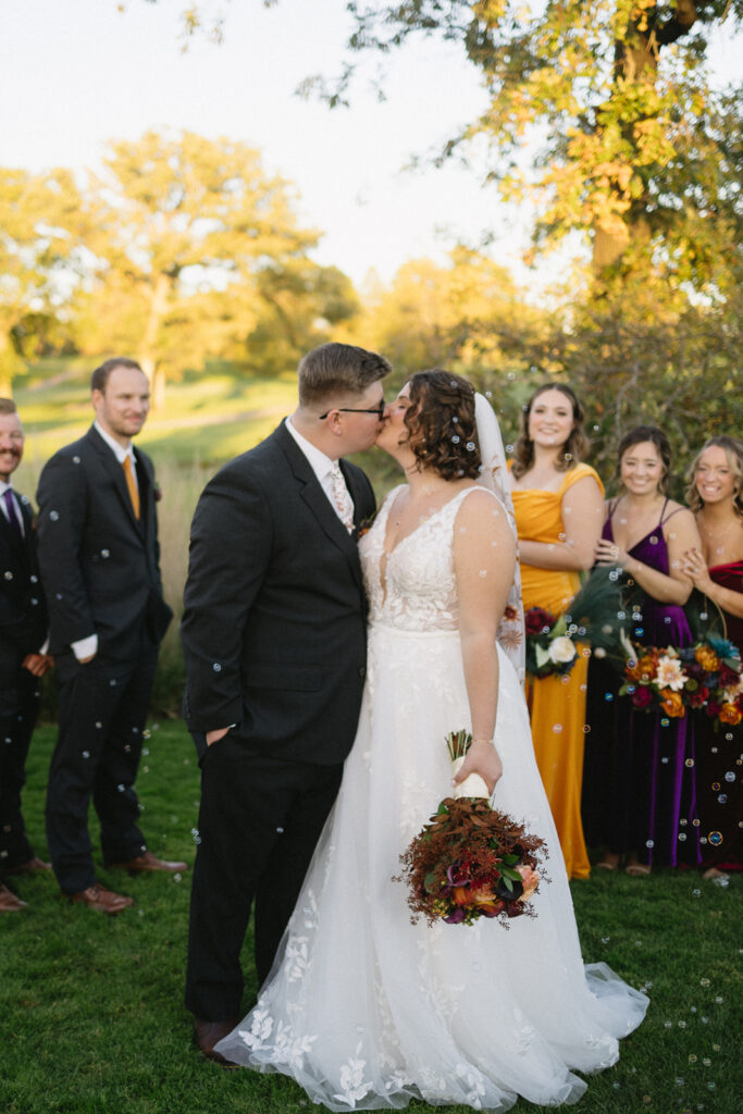 A bride and groom in wedding attire share a kiss outdoors. The bride holds a bouquet. Bridesmaids in colorful dresses and groomsmen stand nearby, smiling. Bubbles float around them, and trees are in the background.