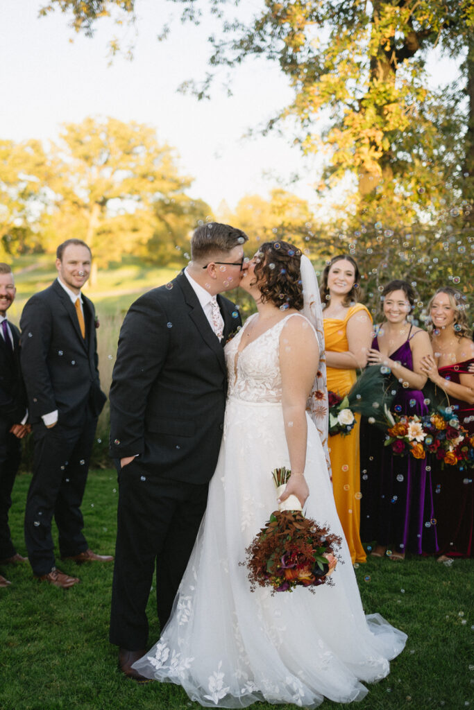 A bride and groom kiss outdoors, surrounded by wedding party members standing under a tree. The bride holds a bouquet, and bubbles float in the air. Bridesmaids are in colorful dresses and groomsmen in dark suits with colored ties.