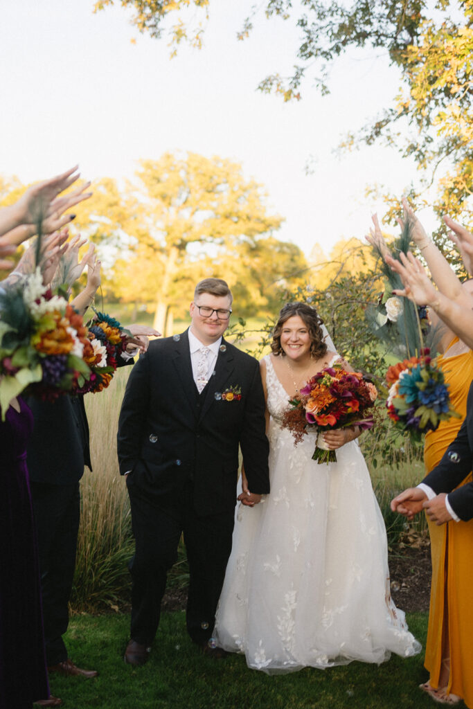 A bride and groom smile while walking under an arch of arms held by guests. The bride holds a vibrant bouquet and wears a lace dress, and the groom is in a black suit with a boutonniere. Sunlight filters through trees behind them, illuminating the path.