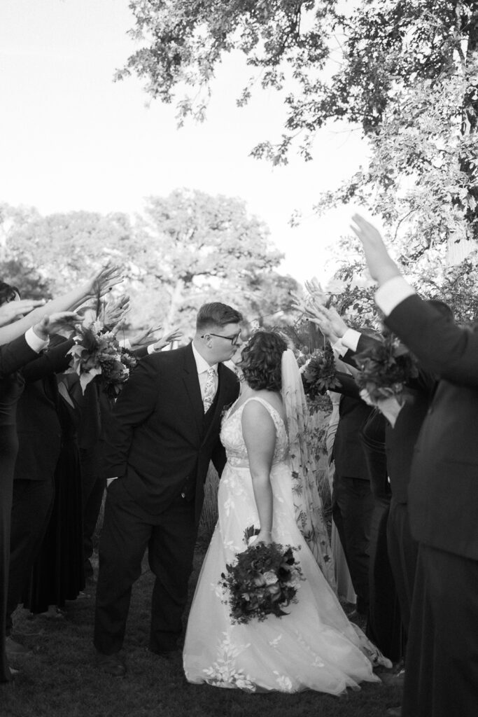 A bride and groom kiss in a grassy outdoor setting. They are surrounded by a joyful group of people holding flowers and raising their hands. The bride wears a gown and veil, and the groom is in a suit. Trees and a clear sky are in the background. Black and white.