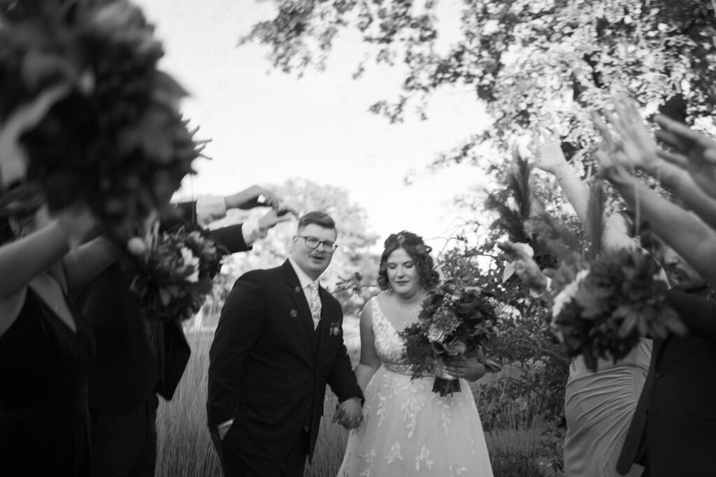 A bride and groom walk hand in hand, smiling, surrounded by people raising bouquets. The couple is dressed in formal attire, and the scene is outdoors with trees and soft lighting. The image is in black and white.