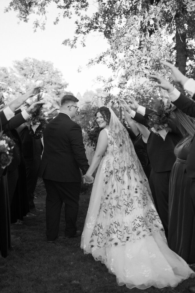 A bride and groom smile while walking hand in hand. The brides floral gown contrasts with the grooms dark suit. Guests on either side hold up bouquets, creating an arch for the couple to walk under. Trees and sunlight are visible in the background.