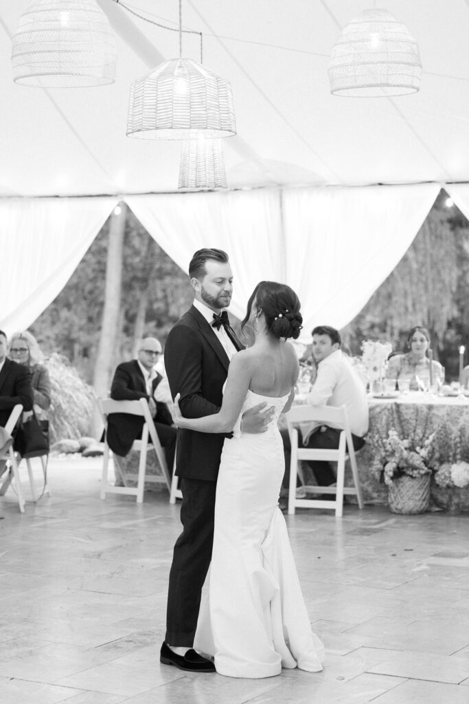 A bride and groom share their first dance under a white tent. The bride wears a strapless gown, and the groom is in a suit. Guests sit at tables in the background, and there are hanging light fixtures above. The scene conveys an elegant wedding celebration.