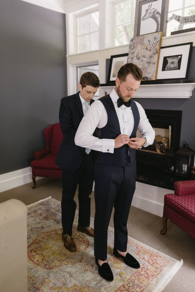 Two men are in a living room. One man is adjusting the others vest. Both are wearing suits, and the man in front also wears a bow tie. The room has white walls, framed art on the mantel, and red armchairs.