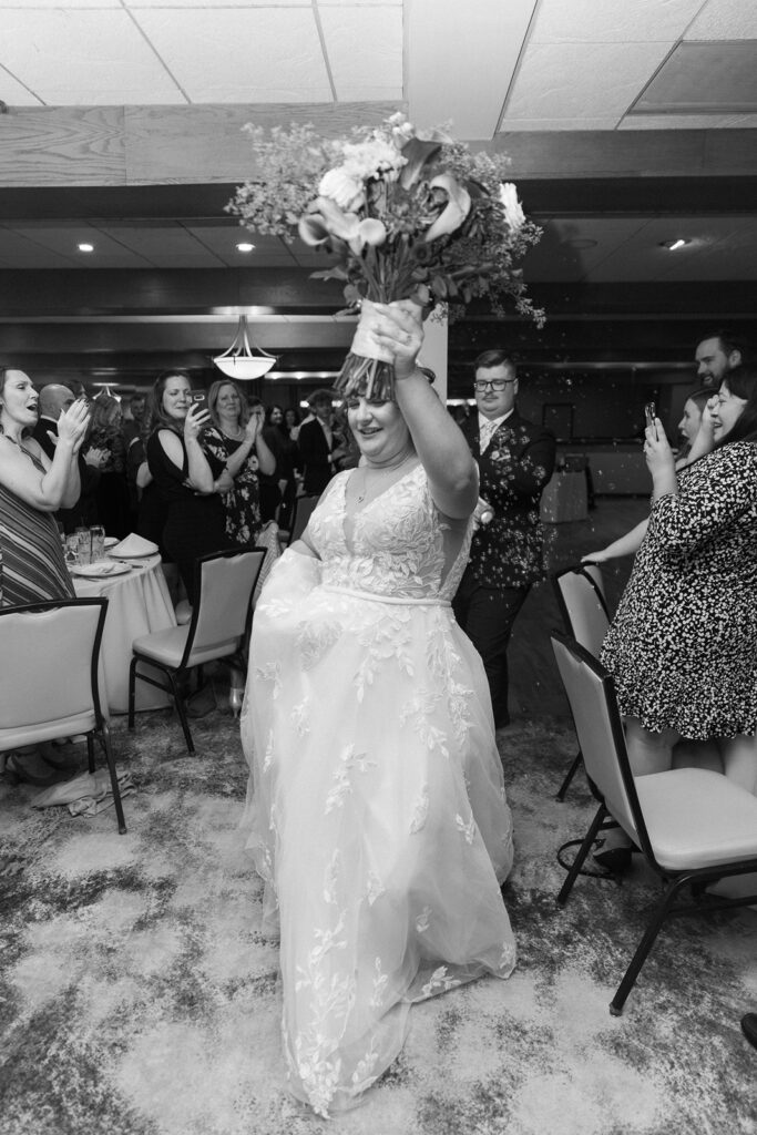 Bride in a lace wedding dress holding a bouquet triumphantly. She walks through a restaurant, surrounded by cheering guests seated at tables. The scene captures joyous celebration and movement.