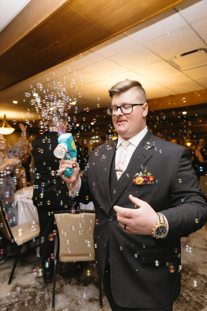 A man in a suit, tie, and glasses holds a bubble machine, releasing bubbles around him. He stands in a lively room with people and chairs visible in the background, against a ceiling with recessed lighting.