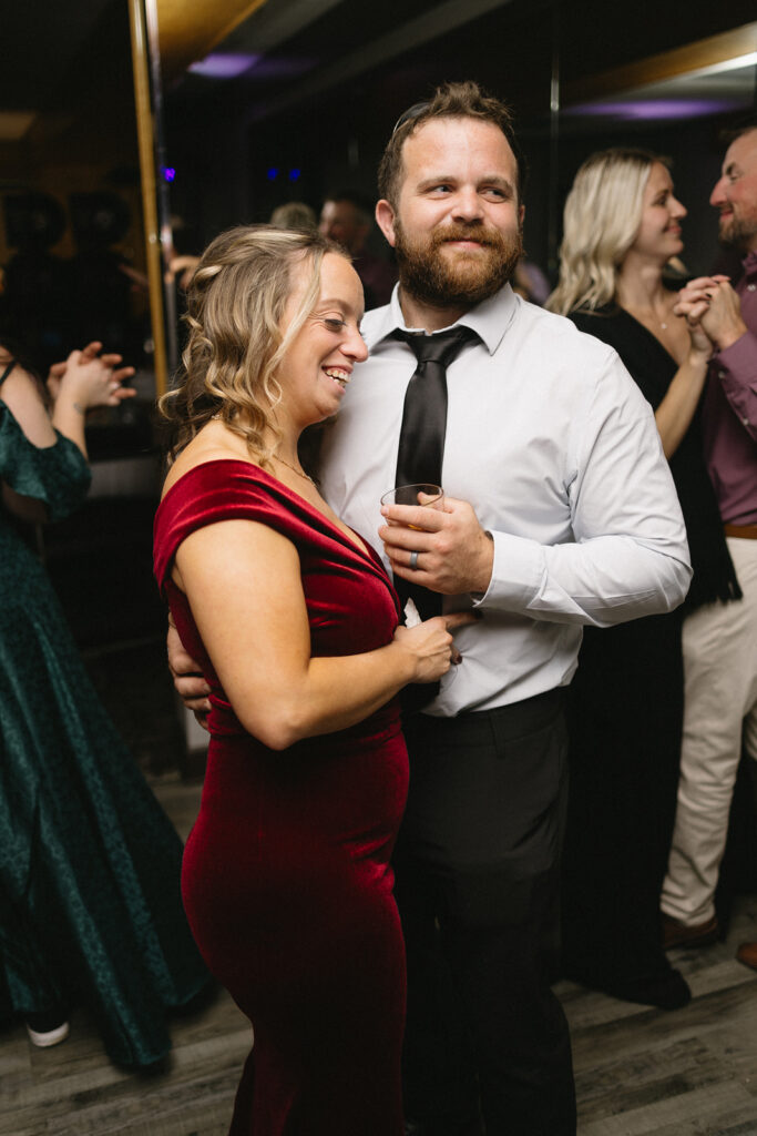 A woman in a red velvet dress and a man in a white shirt and black tie smile while dancing together. Theyre surrounded by other people also dancing in a warmly lit room.