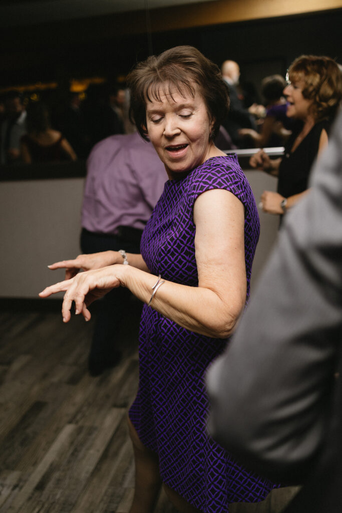 A woman in a purple dress dances at an indoor event. She has short brown hair and is moving her hands rhythmically. Other people are visible in the background, with a dimly lit atmosphere.