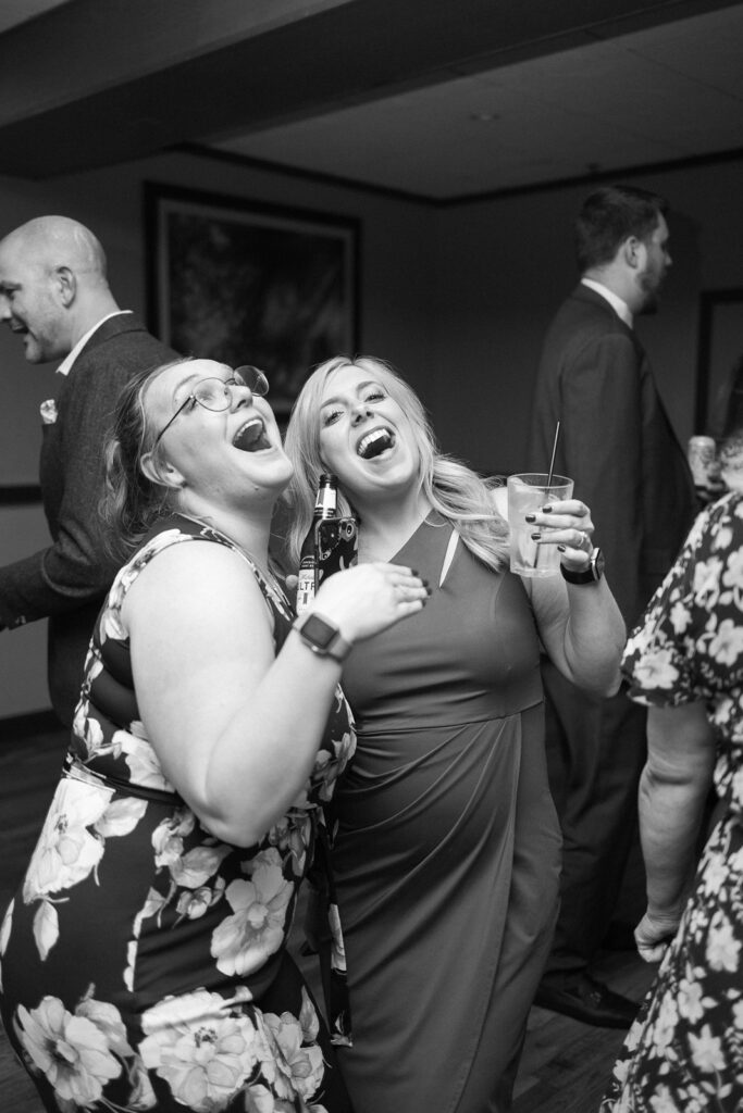 Two women smiling and posing playfully at a social event, one holding a drink and the other with a bottle. They appear to be enjoying themselves. Several people in the background are engaged in conversation. The image is in black and white.