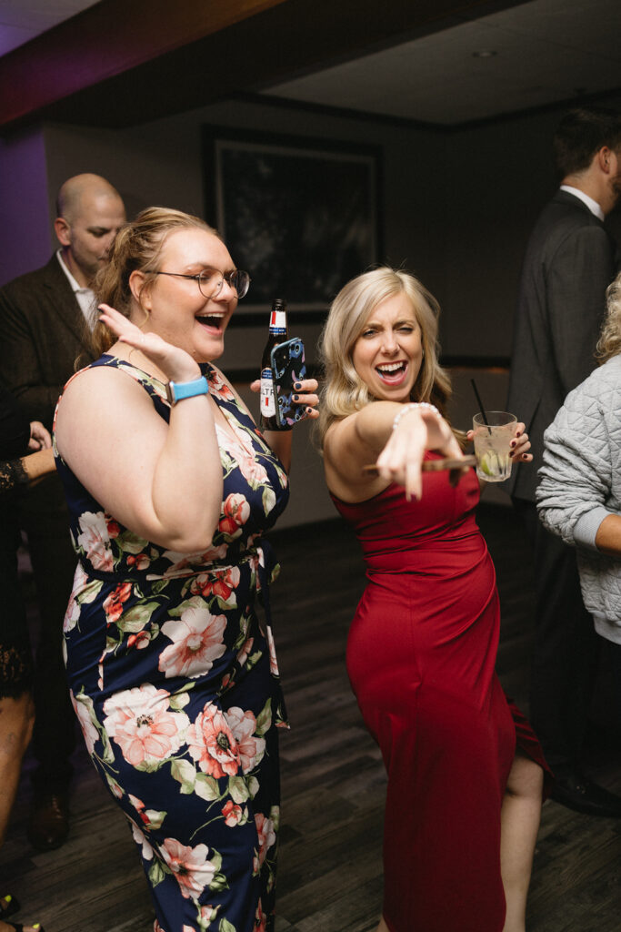 Two women smiling and dancing at a party, one in a floral dress holding a beer and gesturing with her hand, the other in a red dress holding a cocktail and pointing forward. People in the background are socializing. The setting appears to be indoors.