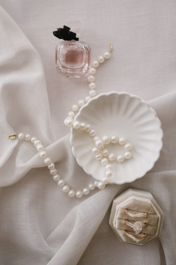 A strand of pearls, a scalloped white dish, a pink perfume bottle, and a beige ring box with three rings are arranged on light, textured fabric—perfect inspiration for Michigan wedding venues or Kalamazoo brides.