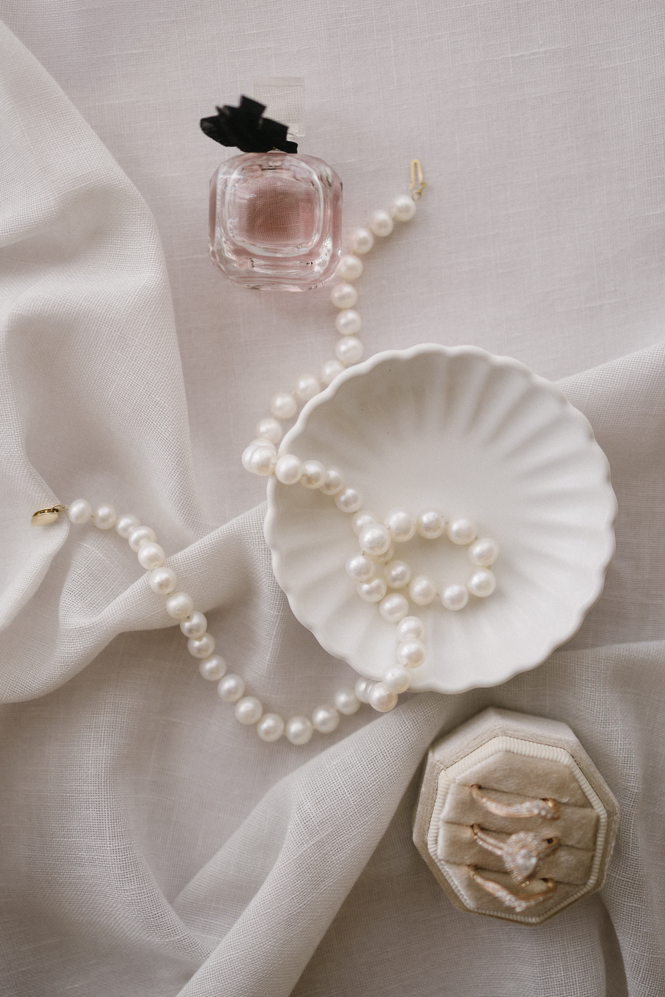 A strand of pearls, a scalloped white dish, a pink perfume bottle, and a beige ring box with three rings are arranged on light, textured fabric—perfect inspiration for Michigan wedding venues or Kalamazoo brides.
