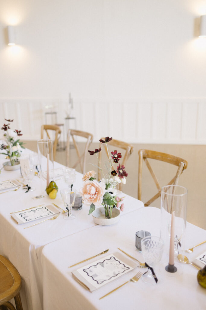 A white table set for a formal event at one of the elegant Kalamazoo wedding venues features gold cutlery, glassware, candles, and a floral centerpiece; wooden chairs line the table in a bright, minimal room in Michigan.