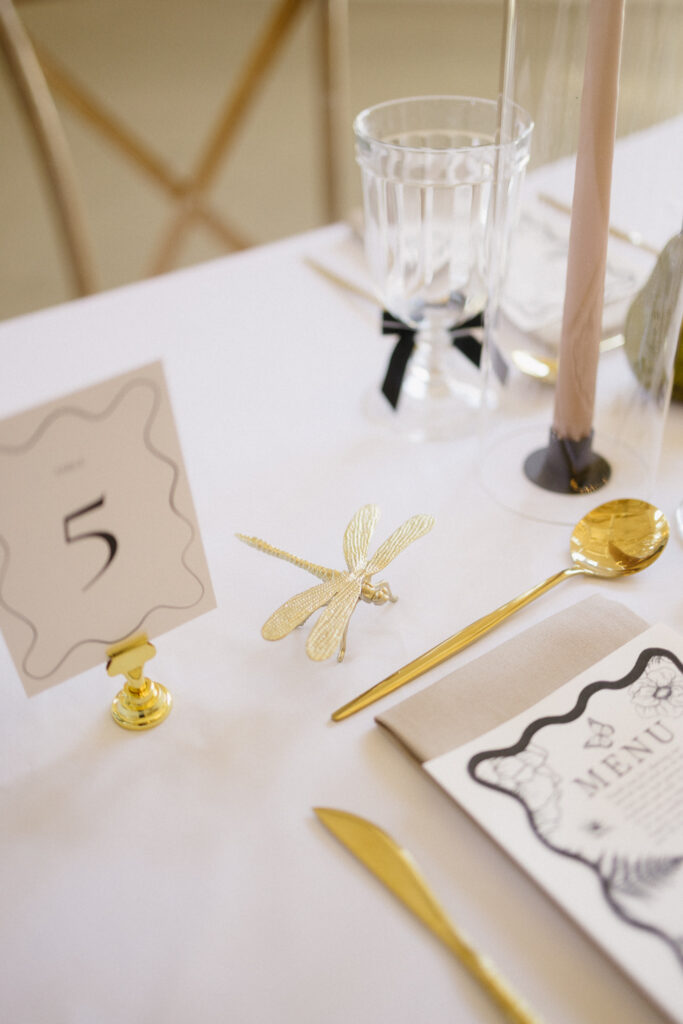 A close-up of an elegant table setting at one of the top Michigan wedding venues, featuring a gold dragonfly decoration, gold cutlery, a menu, a glass, a pink candle, and a table number card displaying the number 5 on a white tablecloth.